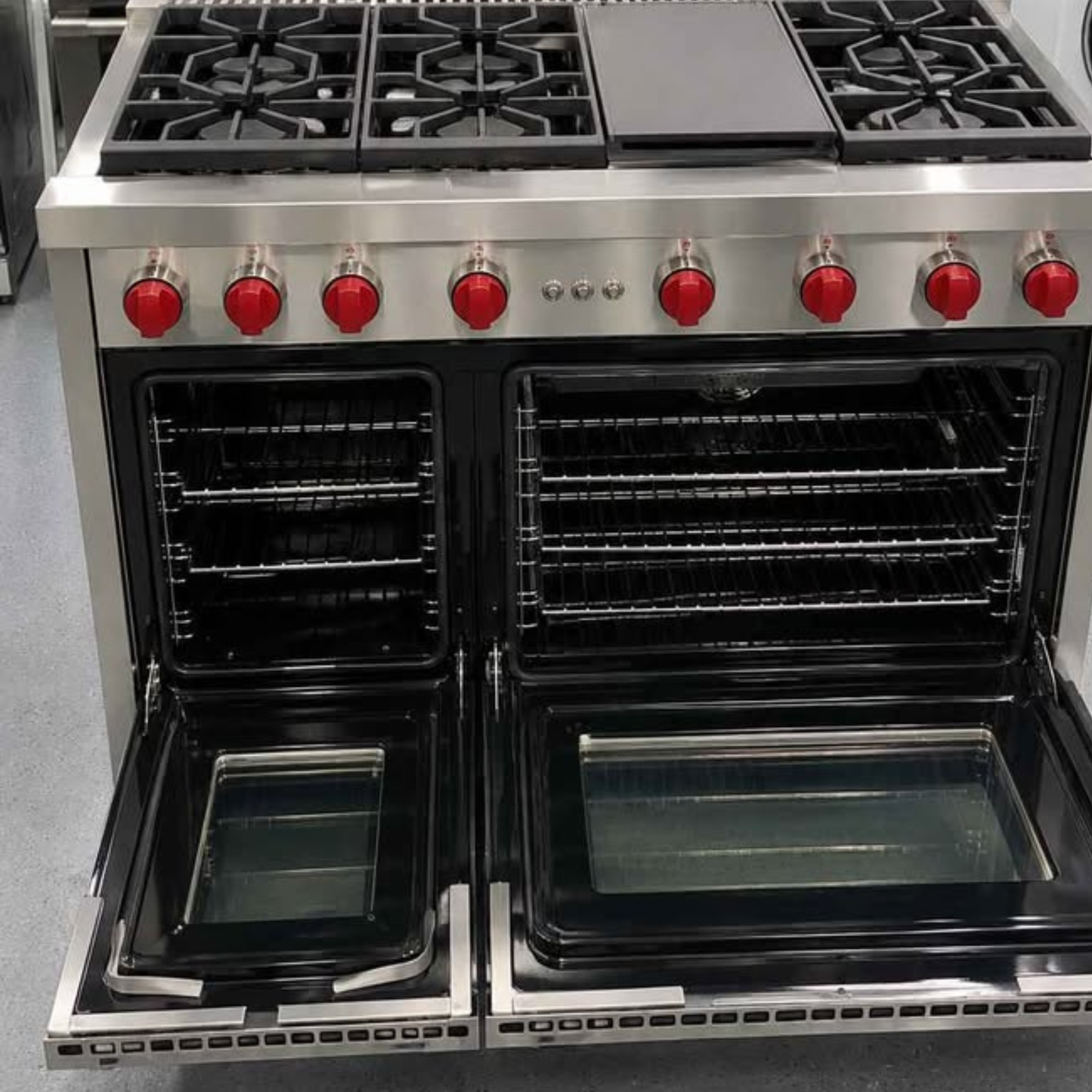A large stainless steel freestanding gas range with two open ovens, black grates, a smooth cooktop, and bright red control knobs, photographed in a showroom setting.