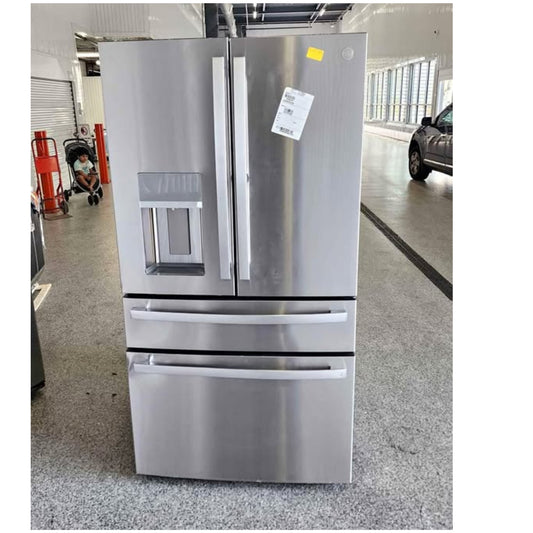 A stainless steel French door refrigerator with two upper doors, a water and ice dispenser on the left door, and two pull-out freezer drawers below, photographed in a warehouse or showroom setting.