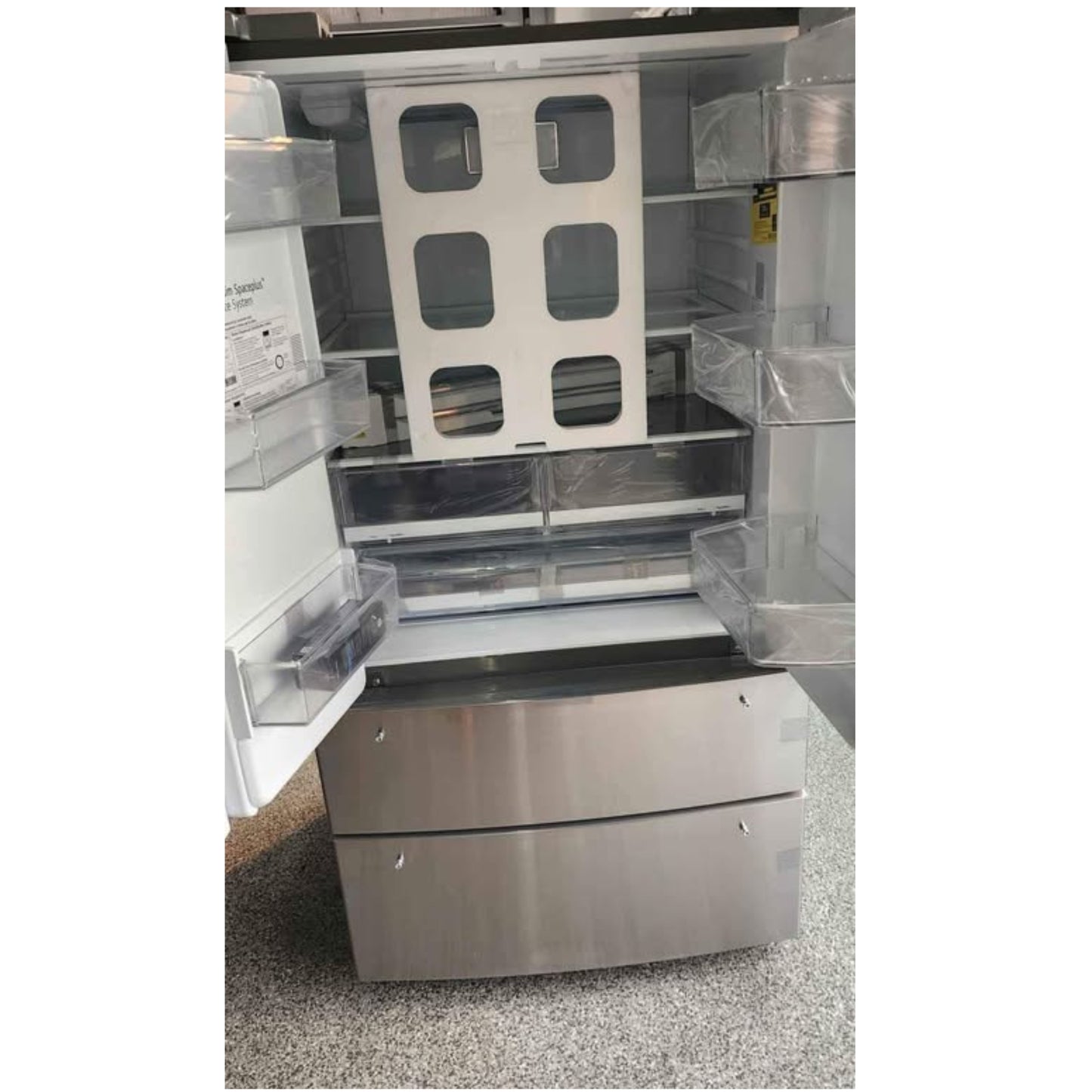 A stainless steel refrigerator shown with its doors open, revealing empty shelves, door bins, and a lower drawer compartment. The appliance is photographed indoors on a speckled floor.